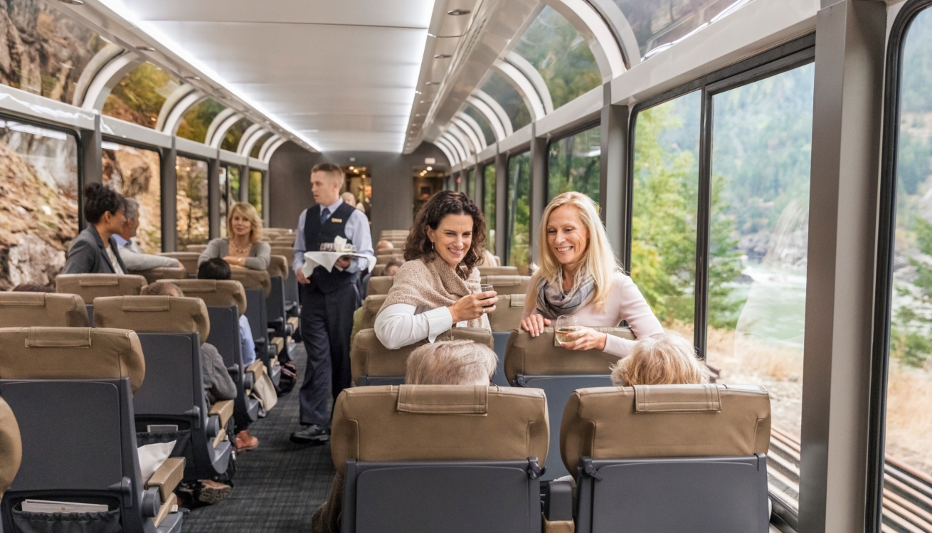 Two women onboard rocky mountaineer train in silverleaf service with wine leaning over chairs